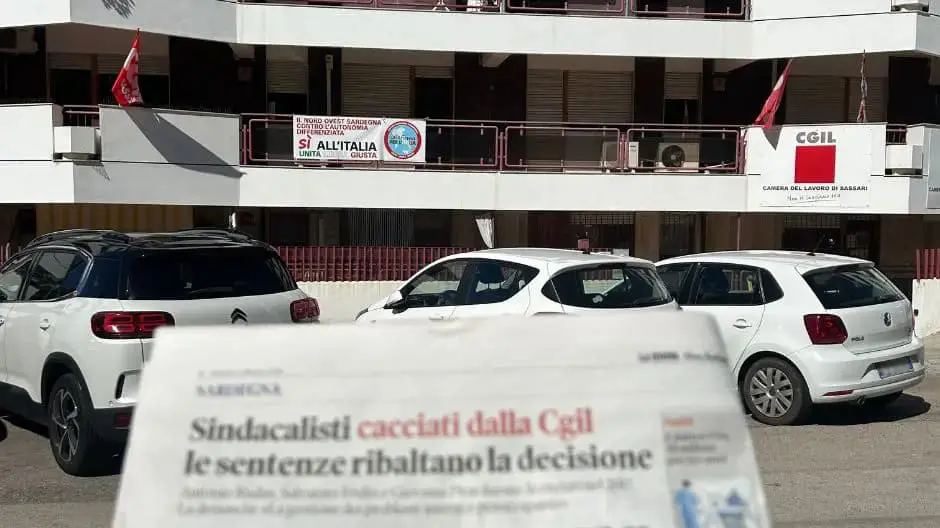 Vista esterna della sede della CGIL di Sassari, con bandiere e cartelli visibili, in primo piano un giornale che titola sulle sentenze che ribaltano i licenziamenti di sindacalisti.